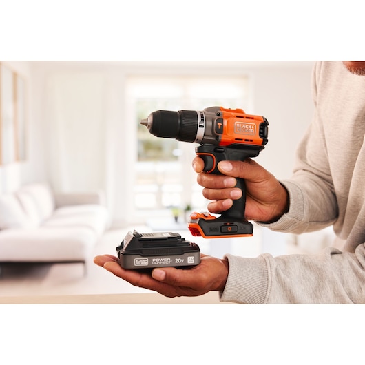 BLACKDECKER BCD383D1 cordless drill and battery held in hand, shown in a bright living room setting.