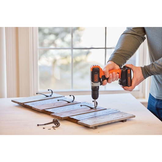 Photo of BLACKDECKER cordless drill BCD70250PK in use, attaching metal hooks to a wooden board on a table, side angle.