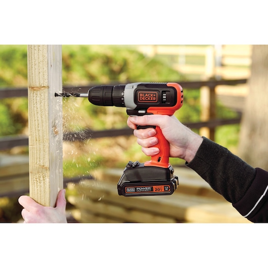 BLACK+DECKER BCD702C1 cordless drill in use, orange and black, drilling into wood outdoors.