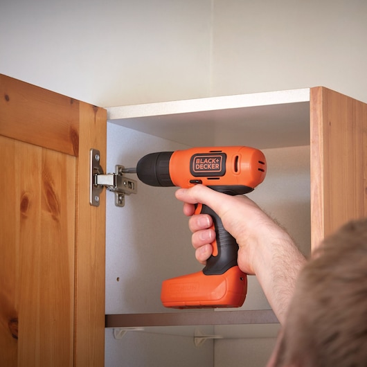 BLACK+DECKER BDCD8HDPK orange cordless drill used to install hinge on wooden cabinet.