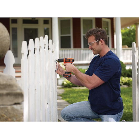 Person using BLACKDECKER BDCDD12C cordless drill to install hinge on white picket fence outdoors.