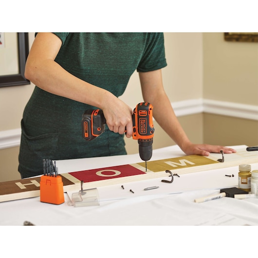 Person using BLACKDECKER BDCDD12C cordless drill on wooden sign at table, drill bits and screws visible.