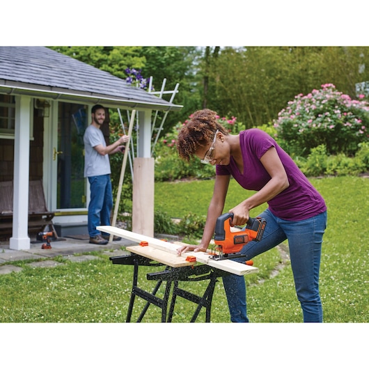 BLACKDECKER BCK22S1S cordless jigsaw used outdoors to cut wood on a workbench in a garden setting.
