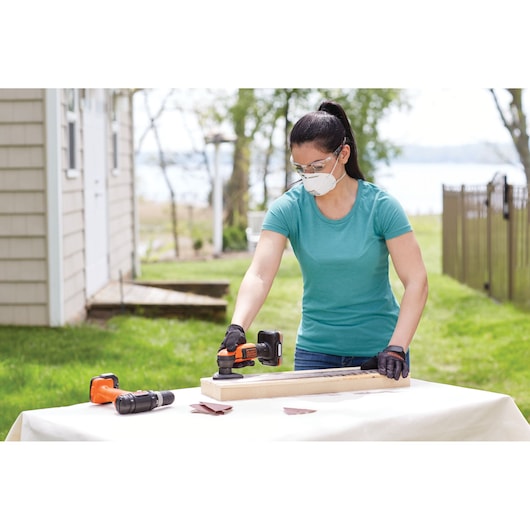 A person using a BLACK+DECKER cordless power tool on a piece of wood outdoors, with another power tool and sanding sheets visible on the table. The person's face is blurred for privacy.