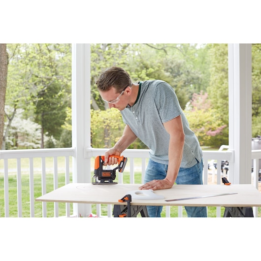 A person using a BLACK+DECKER cordless jigsaw to cut a piece of wood on a table outdoors. The wood is secured with clamps, and the scene shows a porch with greenery in the background. The person's face is blurred.