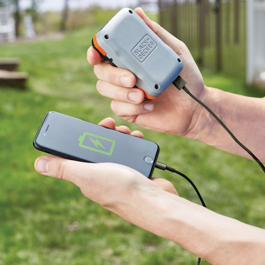 A person holding a BLACK+DECKER portable power bank charging a smartphone outdoors. The phone screen displays a charging battery icon, and both devices are connected with a cable.