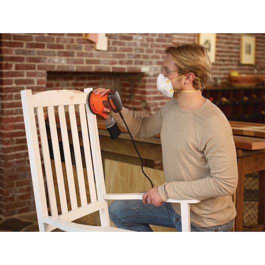 Person using BLACKDECKER BDERO100 orange random orbit sander on white wooden chair indoors.