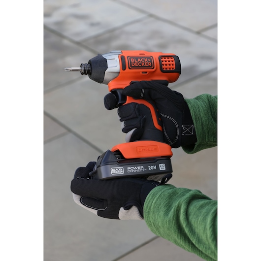 Photo of BLACK+DECKER BDL2020S cordless impact driver, orange and black, held by gloved hands outdoors.