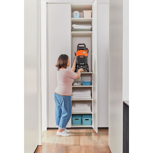 A person places a BLACK+DECKER pressure washer (model BEPW2200) on a shelf inside a white storage closet, surrounded by folded towels and storage bins.