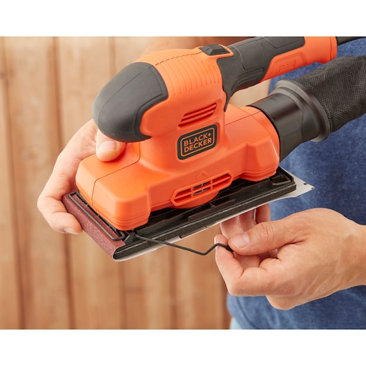 BLACK+DECKER BEW220 orange hand sander shown from side, user adjusting sanding sheet.